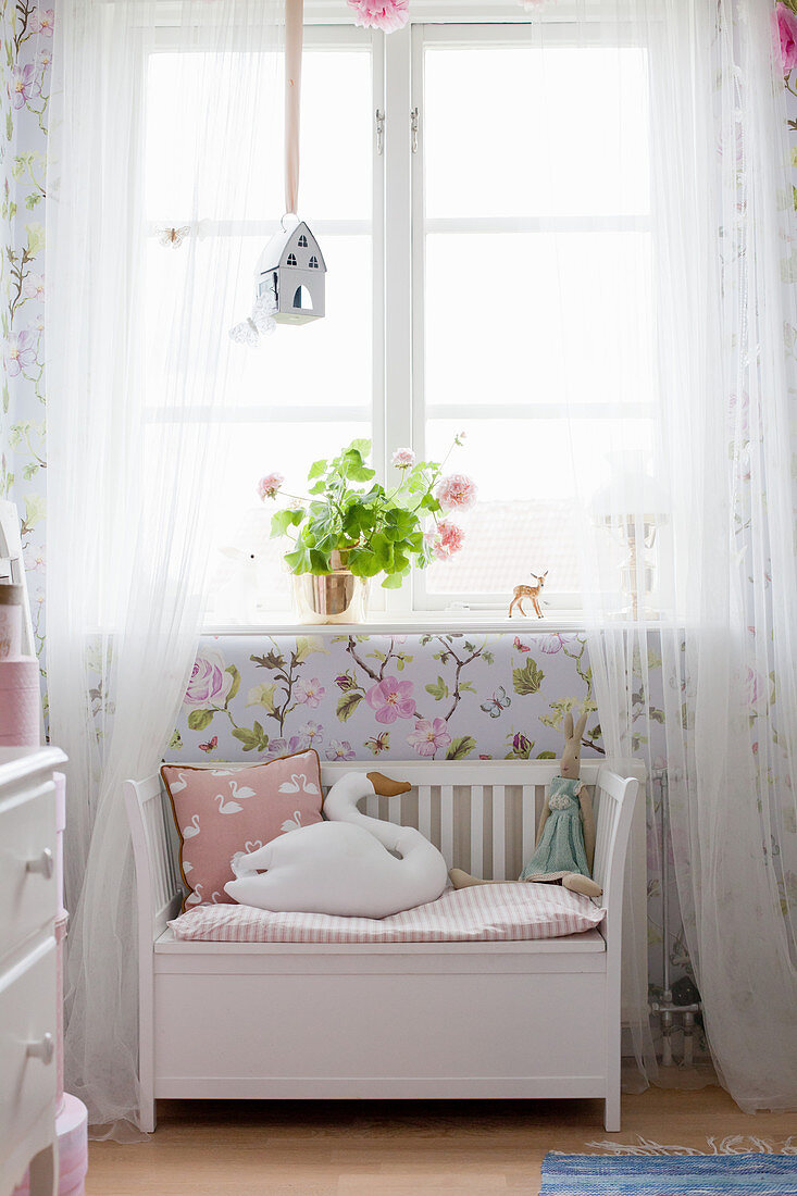 Swan-shaped cushion on white bench below window with airy curtains