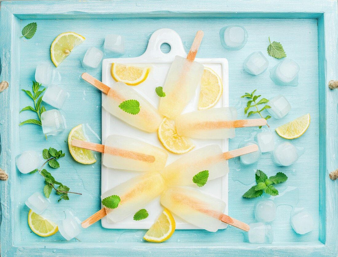 Lemon ice lollies on white ceramic board served with lemon slices, ice cubes and mint leaves