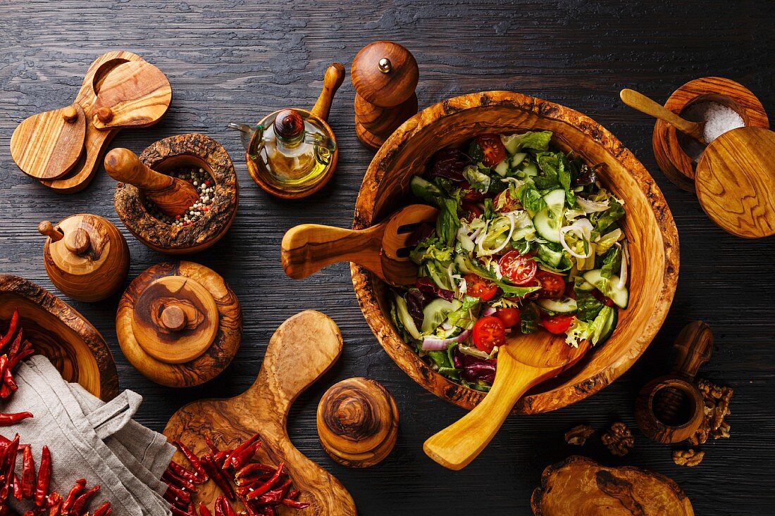 Fresh vegetable salad in olive wood bowl and wooden tableware on black burned wooden background