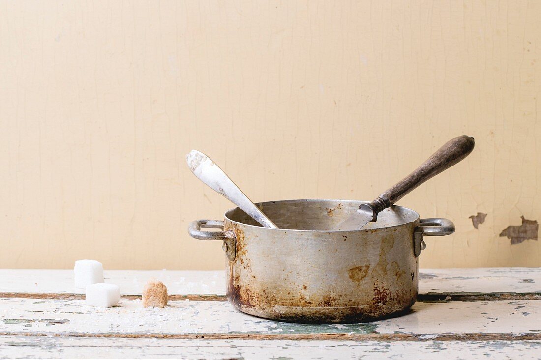 Old aluminum pan of homemade caramel sauce, served with spoons and sugar cubes over white wooden table