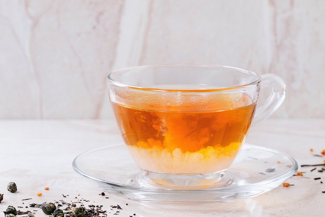 Milk dissolves in glass cup of hot tea on saucer with dry green and black tea leaves