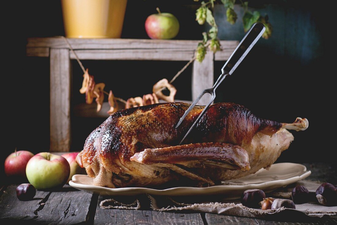 Roast stuffed goose with meat fork in on ceramic plate with ripe apples over wooden kitchen table