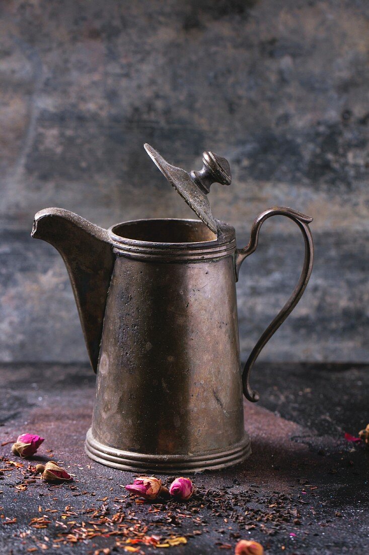 Vintage-Teekanne aus Metall, davor Teeblätter und Rosenknospen
