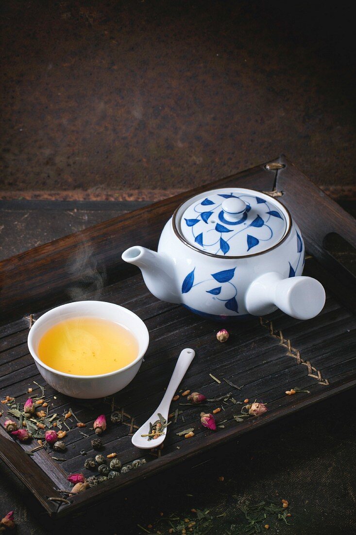 Traditional japanese teapot and cup of green tea, served on bamboo tray with dry tea variations over dark background