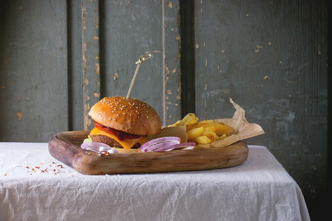Wooden plate with fresh homemade burger and grilled potatoes over white tablecloth with gray wooden background