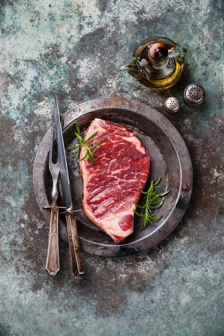 Rohes Striploin Steak mit Rosmarin und Vintage-Besteck auf Teller