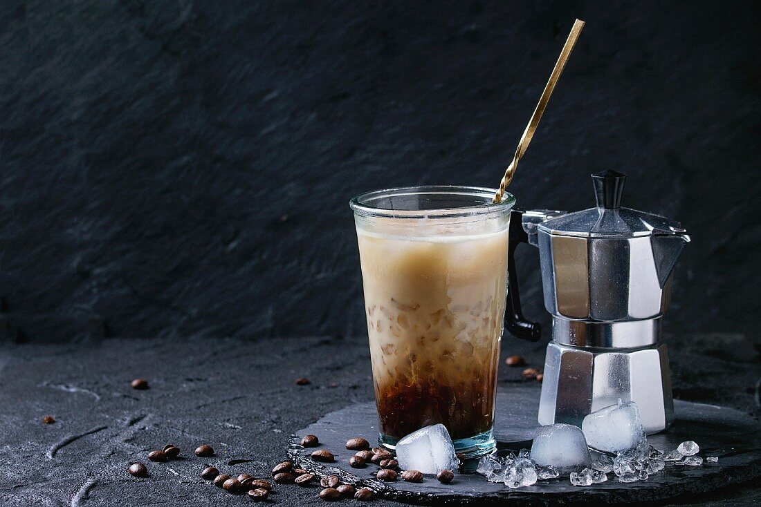 Glass of ice coffee with cream and milk, served with coffee beans, ice cubes and coffee pot on slate stone board
