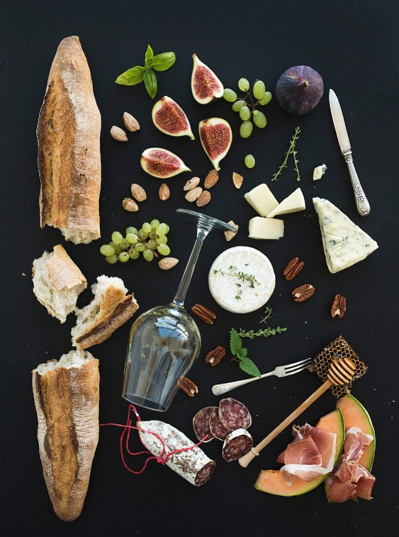 Baguette, glass of white, figs, grapes, nuts, cheese variety, meat appetizers and herbs on black grunge background