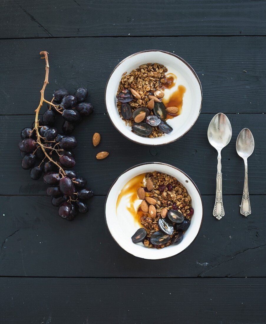 Bowls of oat granola with yogurt, fresh grapes, almond and honey over black wooden backdrop