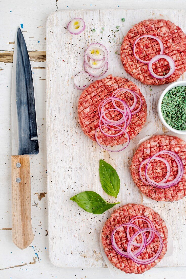 Rohe Burger Patties mit Zwiebelringen und Gewürzen auf weißem Brett