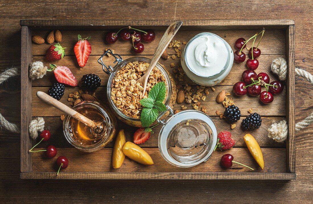 Healthy breakfast ingredients. Oat granola in open glass jar, fruit, yogurt, honey, berries and mint in wooden tray