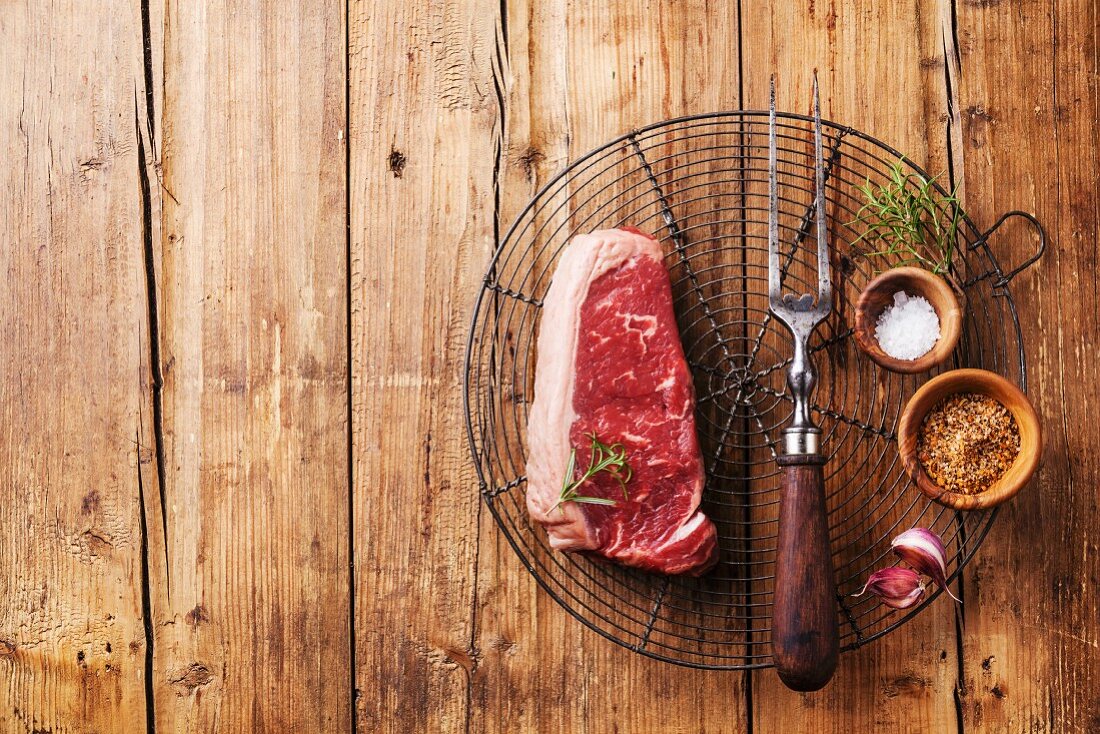 Ein rohes New York Strip Steak mit Fleischgabel und Gewürzen (Aufsicht)