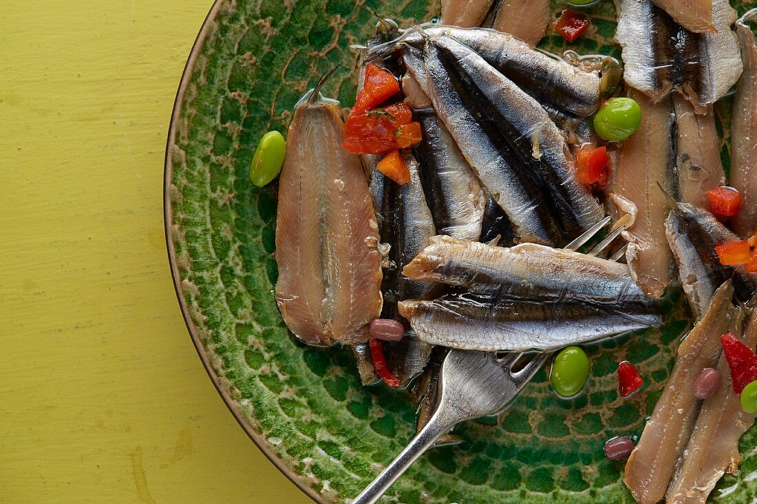 Cold smoked anchovy fillets with edamame and red pepper on rustic green textured plate on vintage yellow table