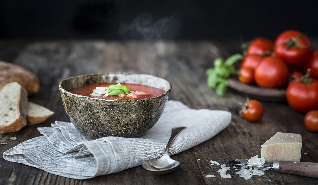 Bowl of steaming tomato soup with fresh tomatoes parmesan cheese bread and basil on a wooden table