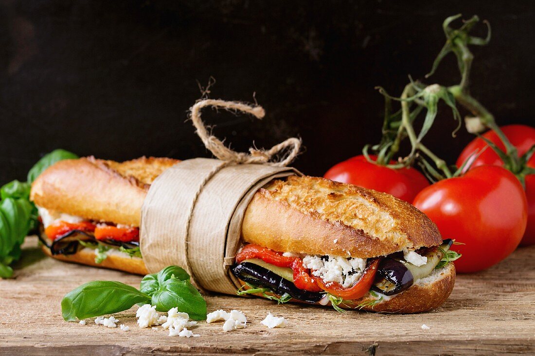 Papered vegetarian baguette submarine sandwich with grilled eggplant, pepper and feta cheese on wooden chopping board
