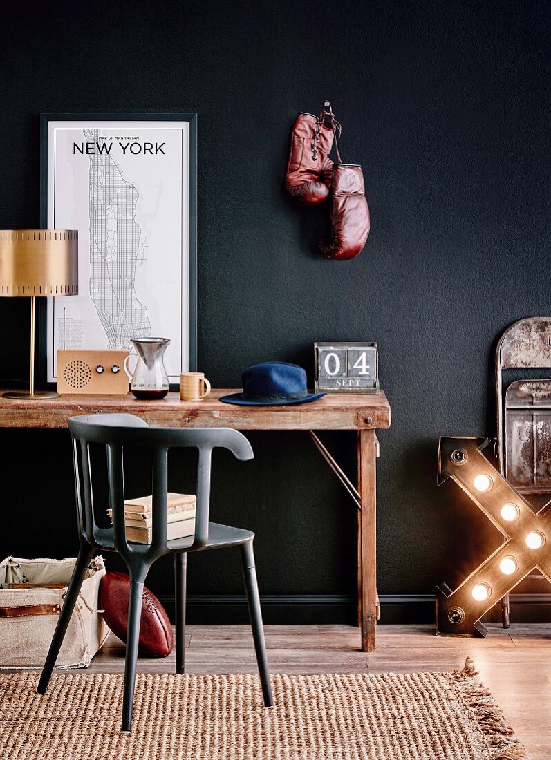 Rustikaler Schreibtisch mit Messing-Tischleuchte, New-York-Karte, Radio, Kaffeekanne und Hut vor schwarzer Wand mit Boxhandschuhen