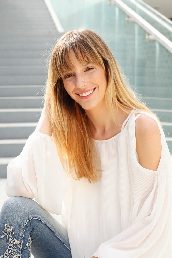 Blonde Frau in weißer Bluse und Jeans mit Blumenstickerei sitzt auf einer Treppe