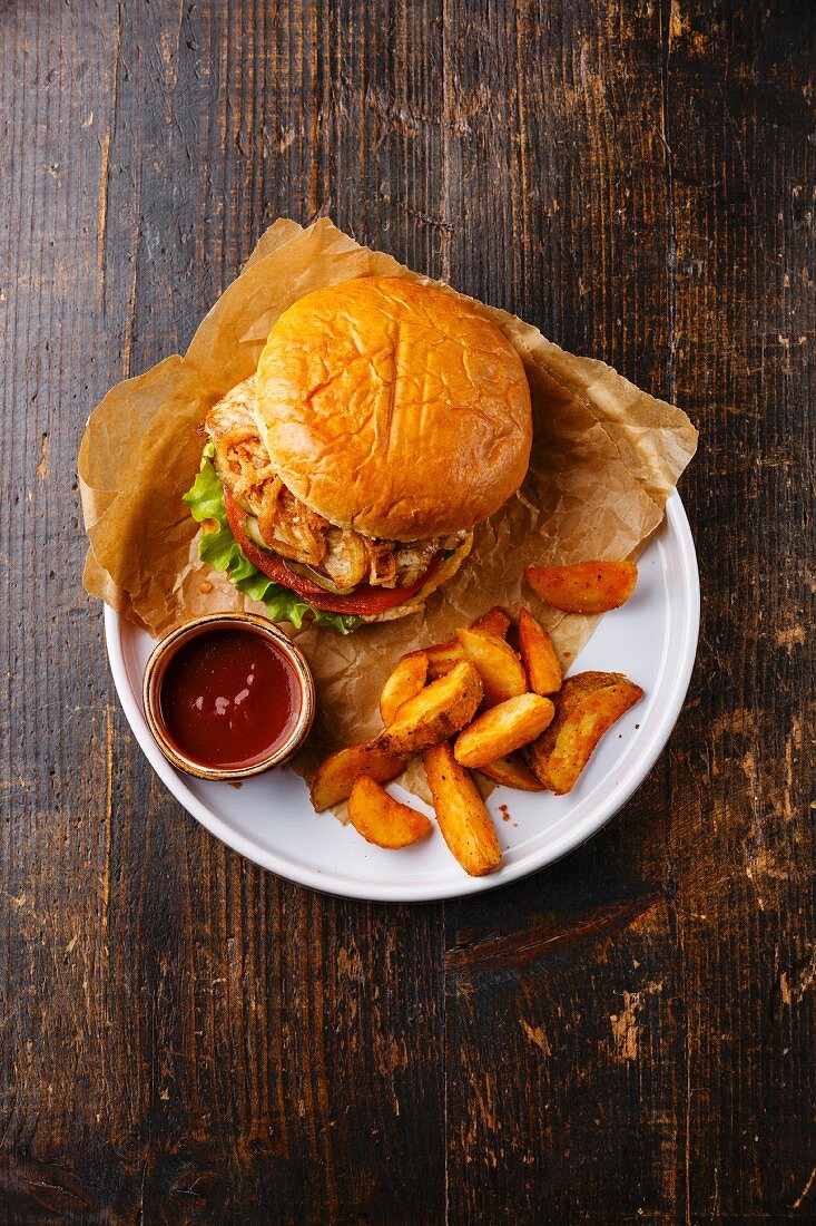 Burger with chicken breast and fried onions with potato wedges on plate on dark wooden background