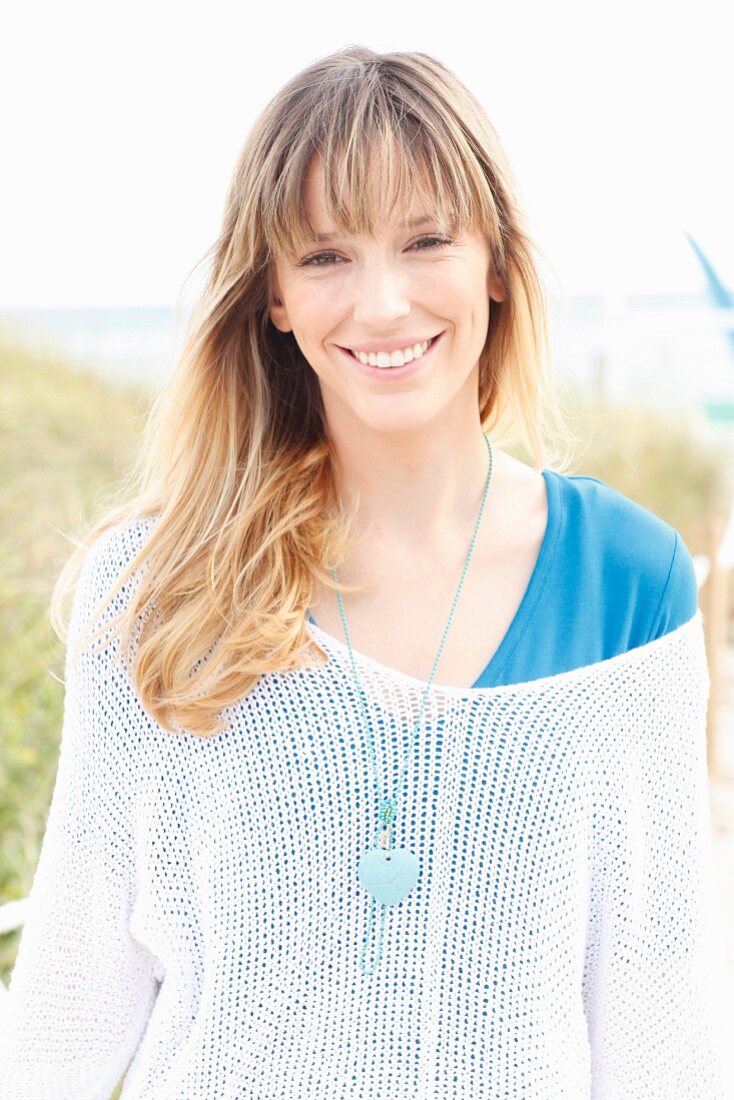 A blonde woman wearing a blue T-shirt, a white knitted jumper and a necklace on the beach
