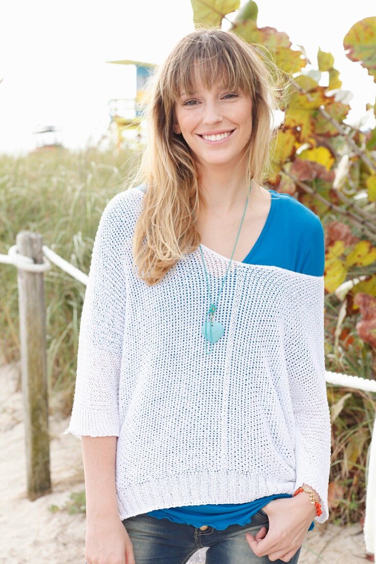 A blonde woman wearing a blue T-shirt, a white knitted jumper and jeans on the beach