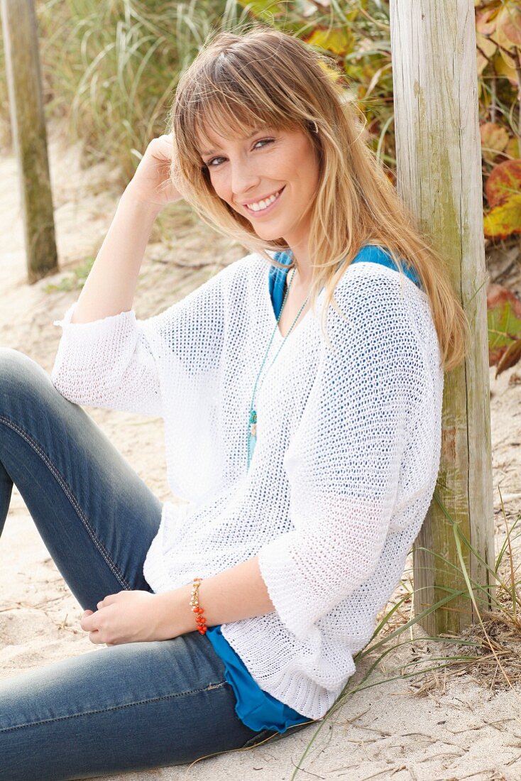 Blonde Frau in blauem T-Shirt, weißem Strickpullover und Jeans am Strand