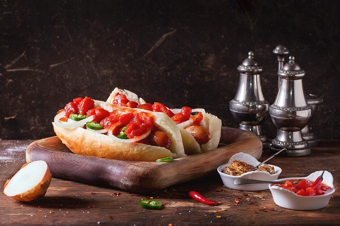 Homemade hot dogs on wooden plate with ingredients mustard, tomato sauce, onion, pepper, rosemary