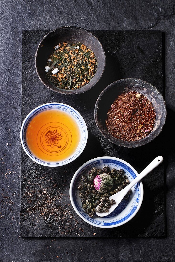 Grüntee, Roibusch, Teeperlen mit Teeblume sowie aufgebrühter Tee in Teeschälchen