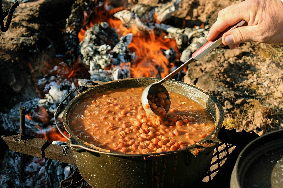 The hand of man stirring beans with a ladle over a campfire