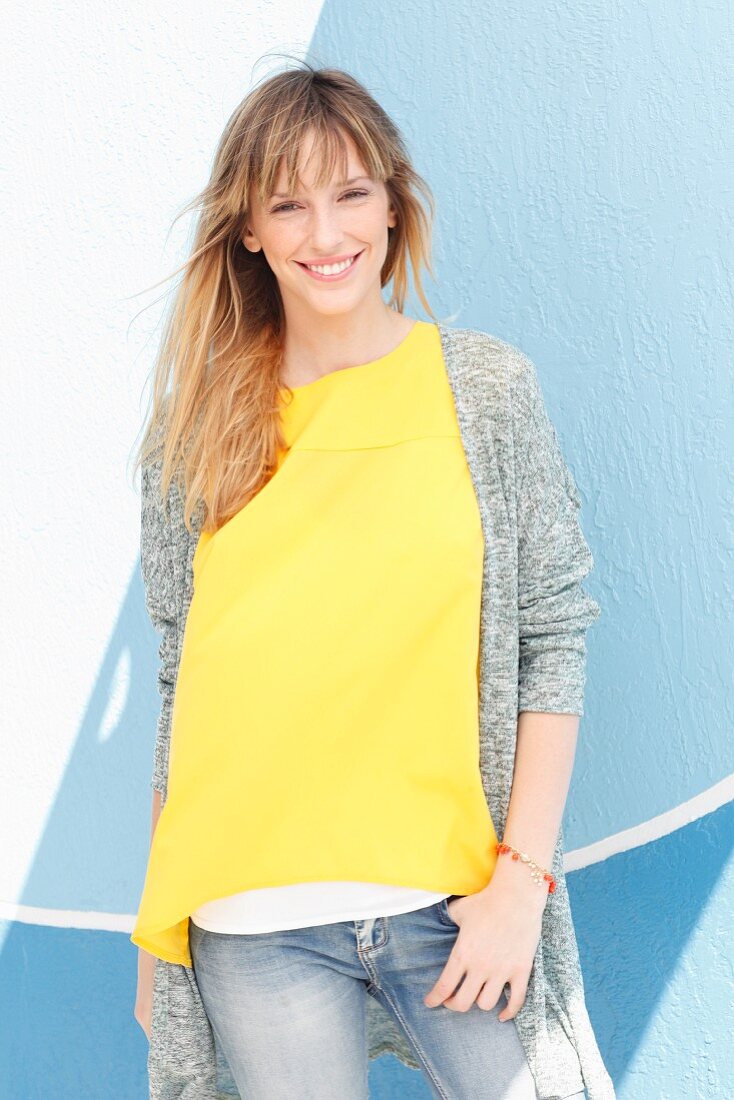 A blonde woman wearing a yellow top, a grey cardigan and jeans in front of a blue wall