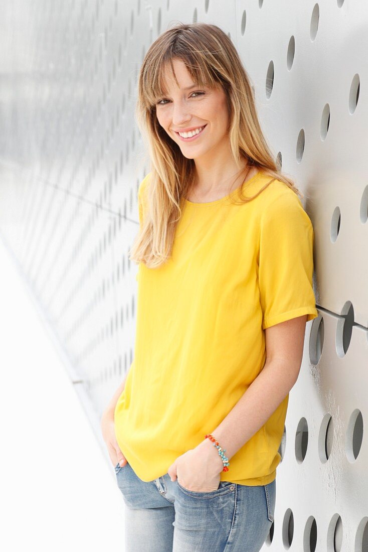 A blonde woman wearing a yellow T-shirt and jeans in front of a wall with holes