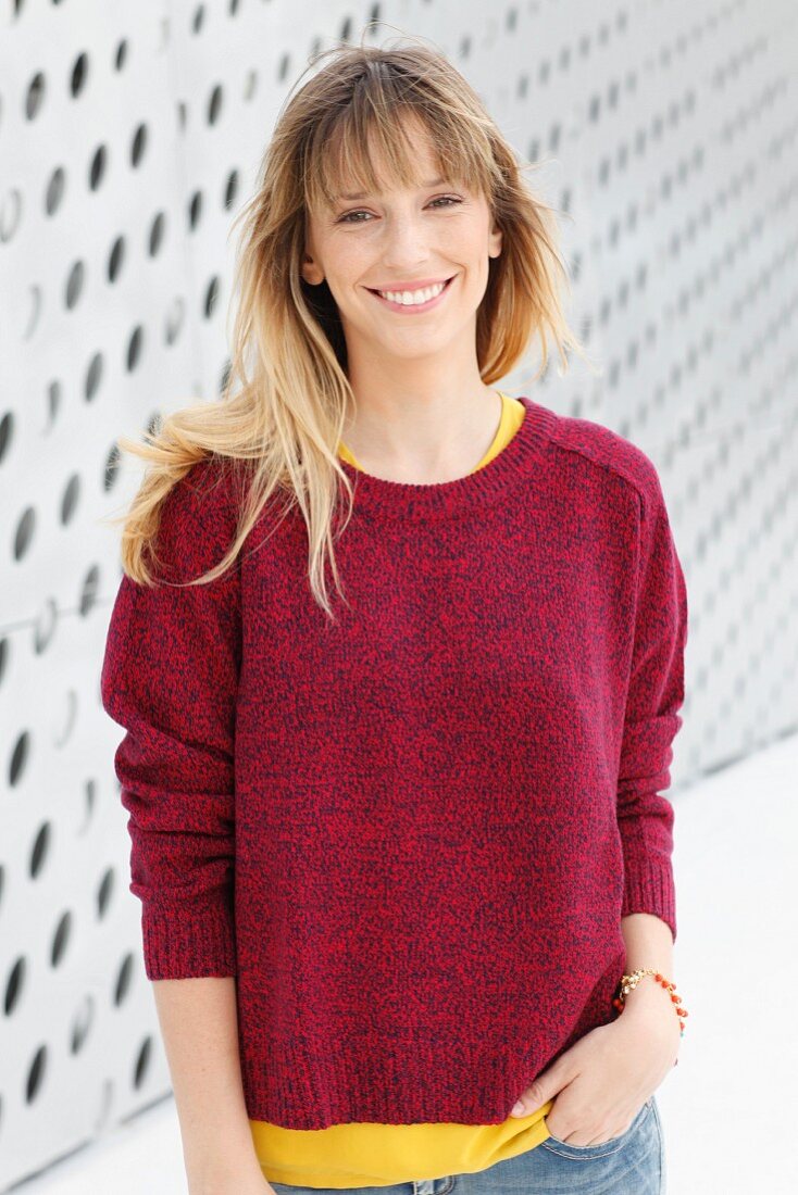 A blonde woman wearing a yellow T-shirt, a red jumper and jeans in front of a wall with holes in it