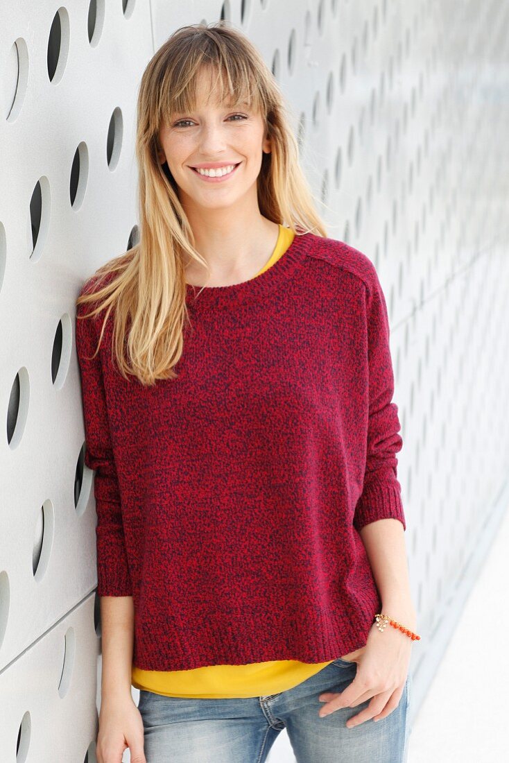 Blonde Frau in gelbem T-Shirt, rotem Strickpullover und Jeans vor Wand mit Löcher