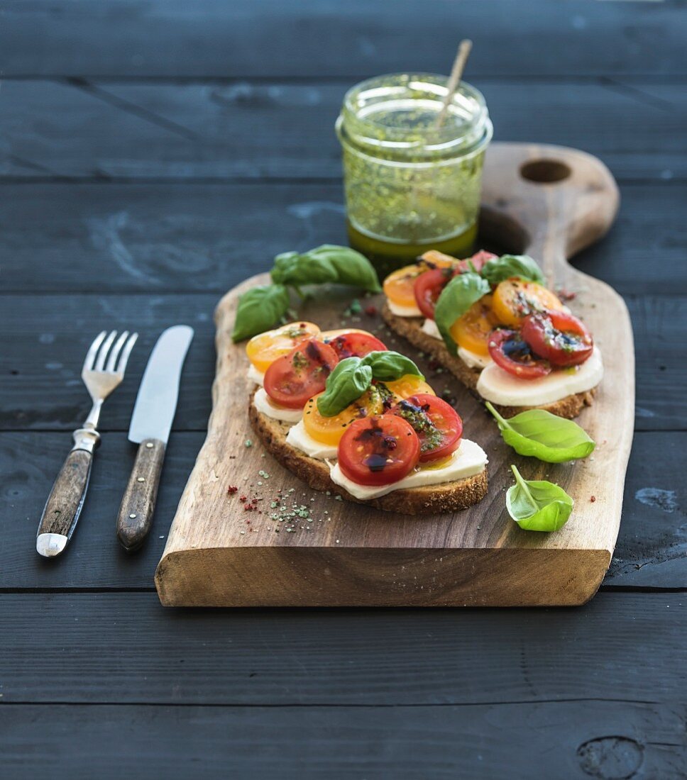 Tomato, mozzarella and basil sandwiches on dark wooden chopping board, pesto jar over black background
