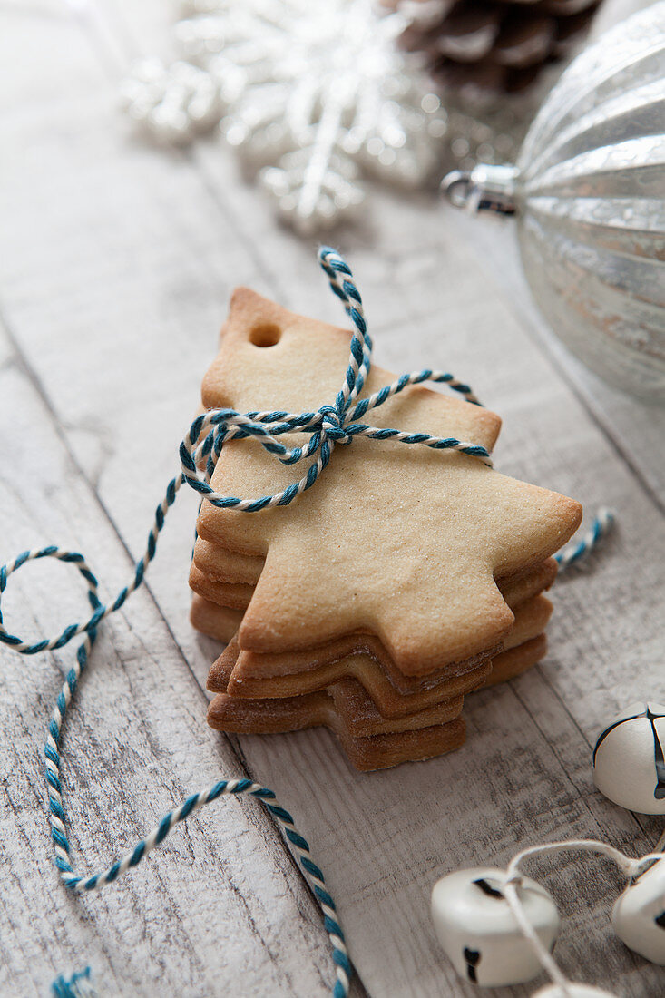 Stack of Christmas tree shaped biscuits tied with blue and white baker twine on a rustic white wood suface and surrounded with Christmas baubles and bells