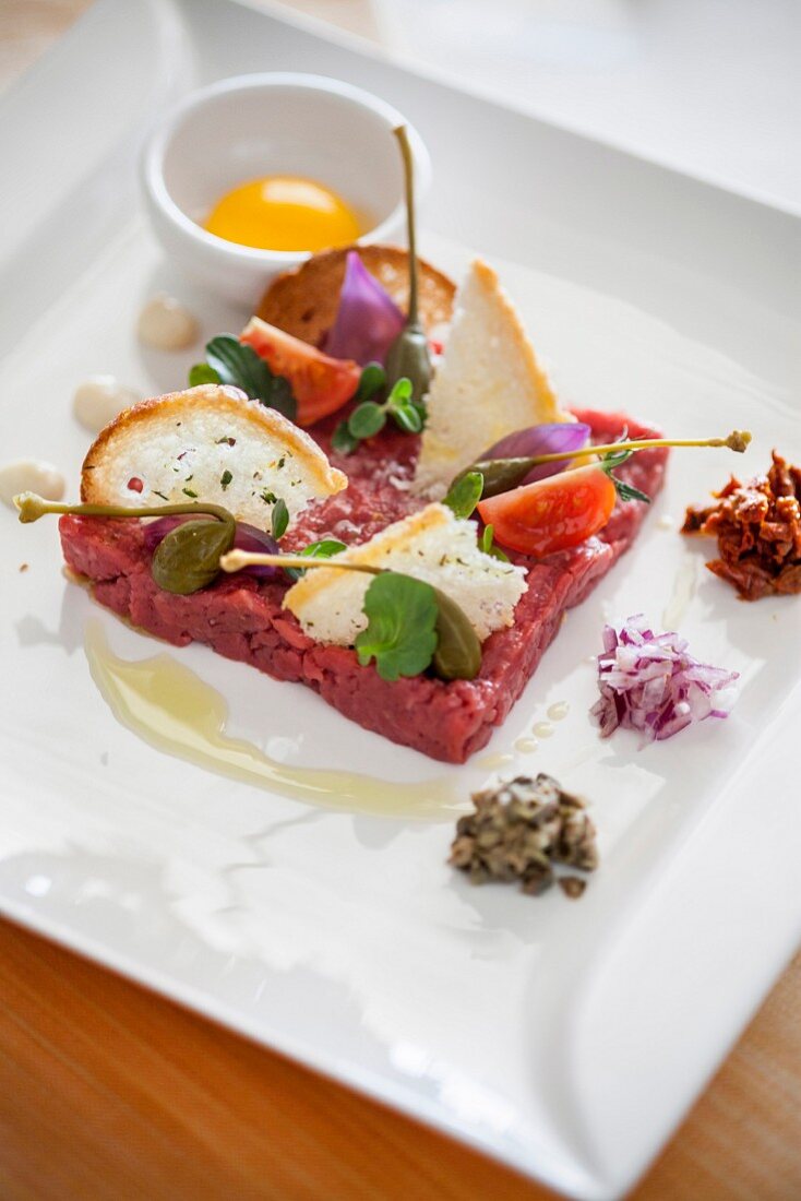 Beef tartare with capers, chopped onion, dried tomatoes in oil and croutons, next to an egg yolk in bowl