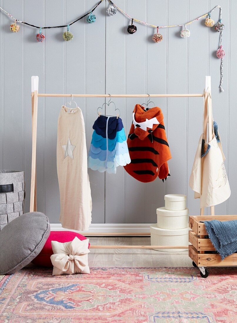 Wooden clothes rack with children's costumes in front of gray wooden paneling and pompom chain