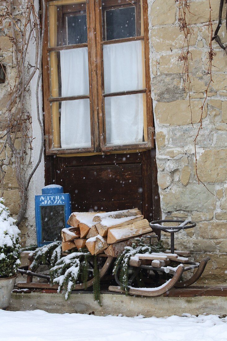 Snowy firewood and pine branches on old wooden sledge against stone façade