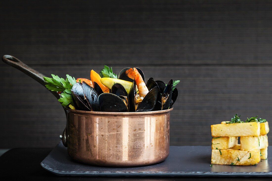 A copper saucepan of mussels with prawns, corn and vegetables served with a stack of chunky chips