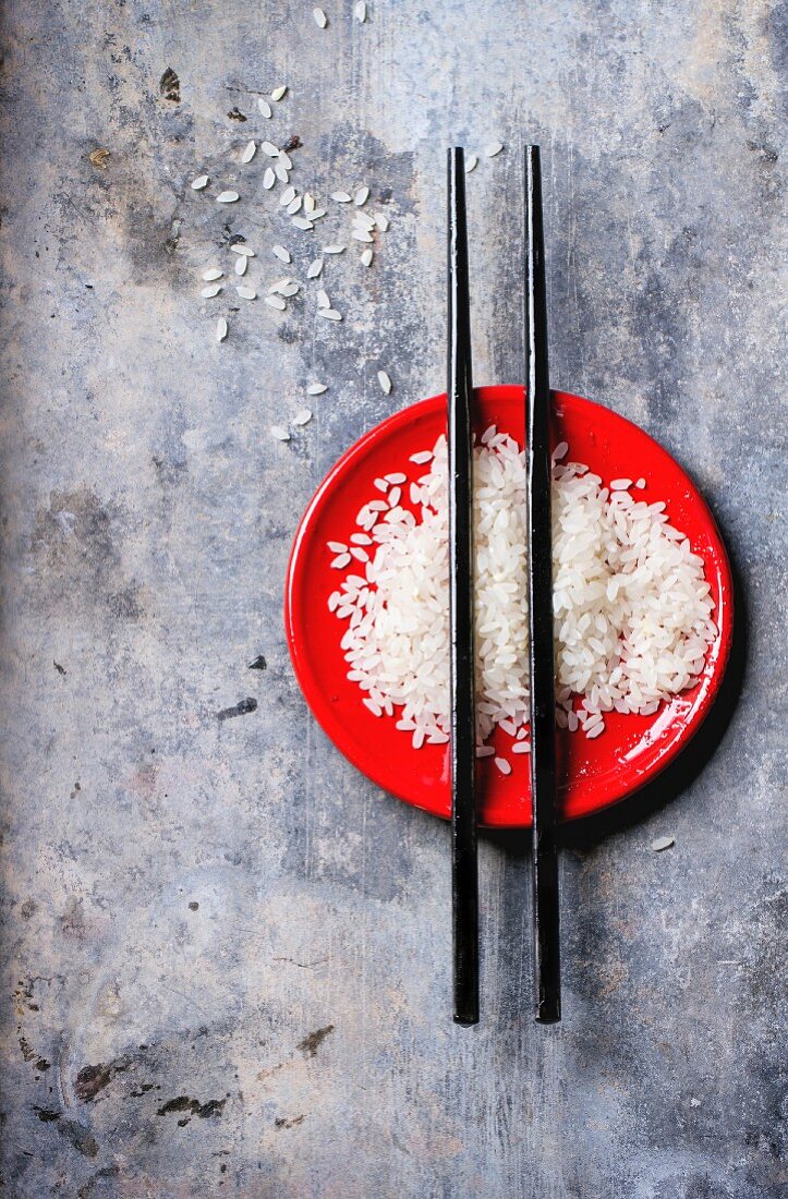 Ungekochter Reis mit Essstäbchen auf rotem Teller vor grauem Hintergrund
