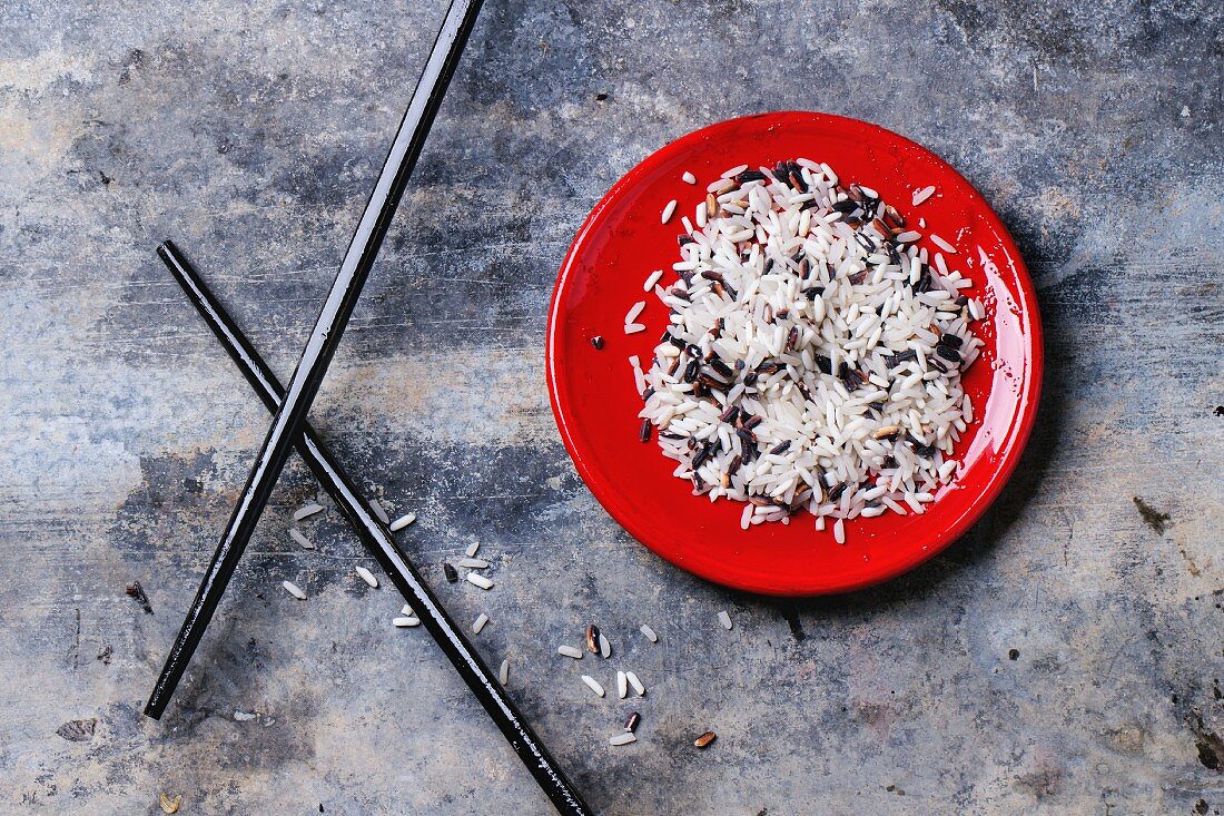 Ungekochter Reis auf rotem Teller, daneben Essstäbchen (Aufsicht)