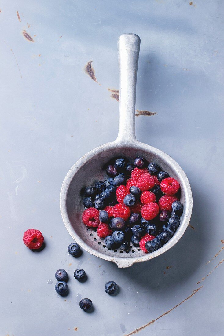 Aluminium colander with fresh raspberries and blueberries over gray metal background