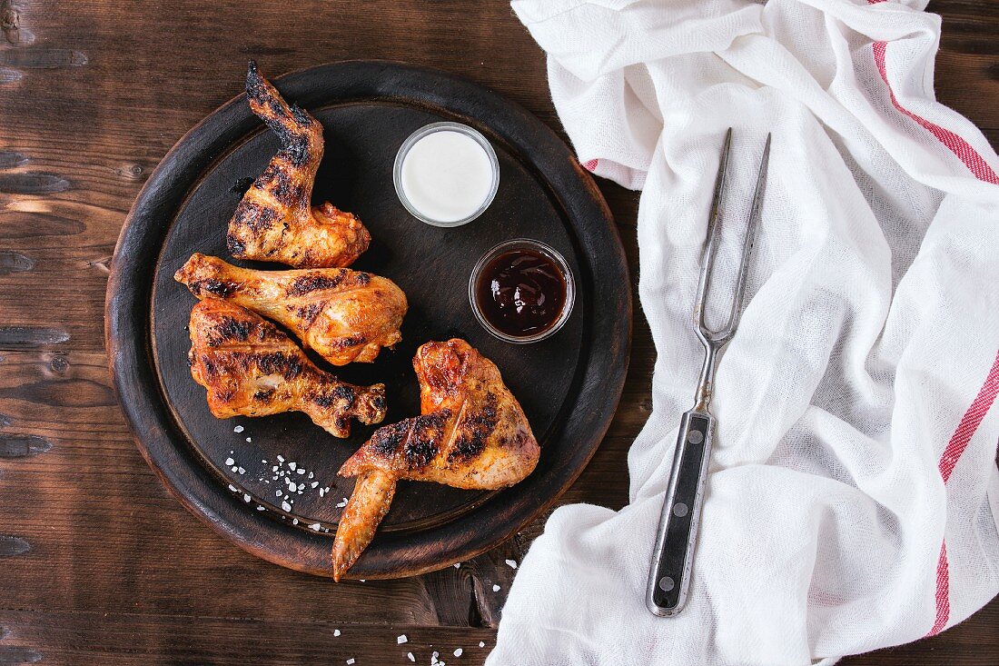 Grilled BBQ chicken meat wings and legs served on round wood chopping board with two sauces and salt