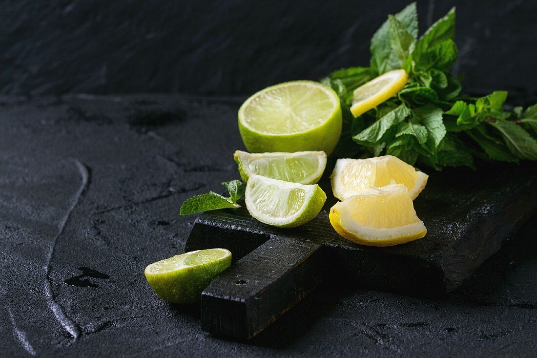 Sliced Lime and lemons with bunch of fresh mint on black wooden chopping board over black textured background