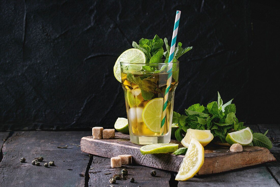 Glass of Iced green tea with lime, lemon, mint and sugar cubes on wooden chopping board