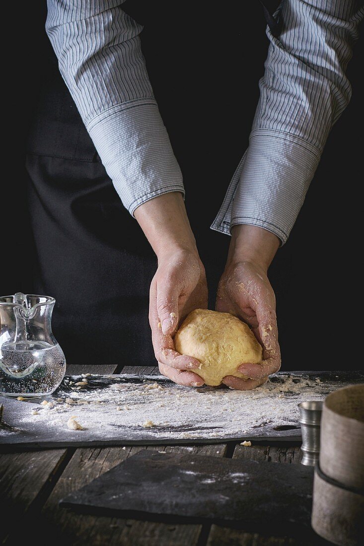 Female hands hold ready-made dough for pasta on old wooden kitchen table, powdered by flour