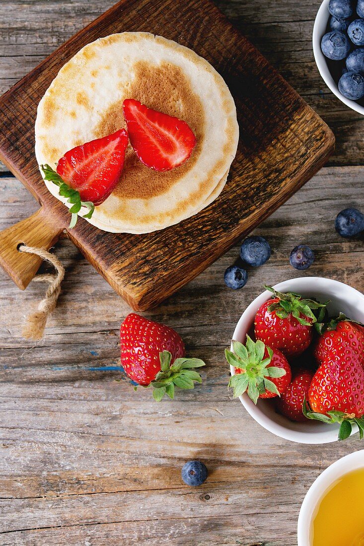 Pancakes with strawberries and blueberries, bowls of honey and fresh berries over old wooden background