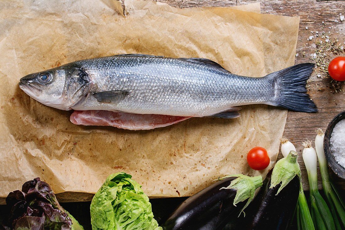 Raw uncooked seebass fish on baking paper with sea salt, dry herbs and vegetables