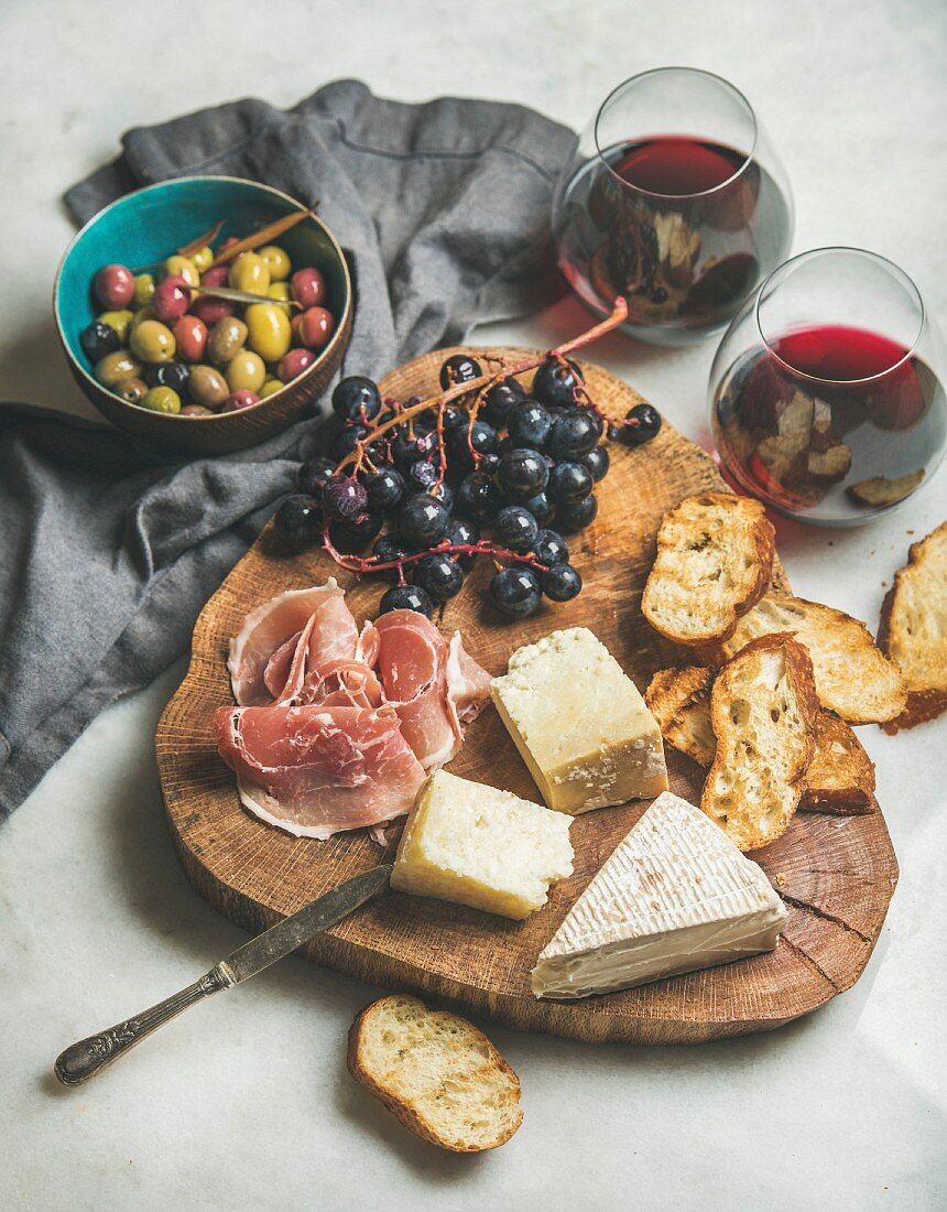 Käse, Schinken, Röstbrot und Trauben auf Holzbrett, dazu Oliven und Rotwein
