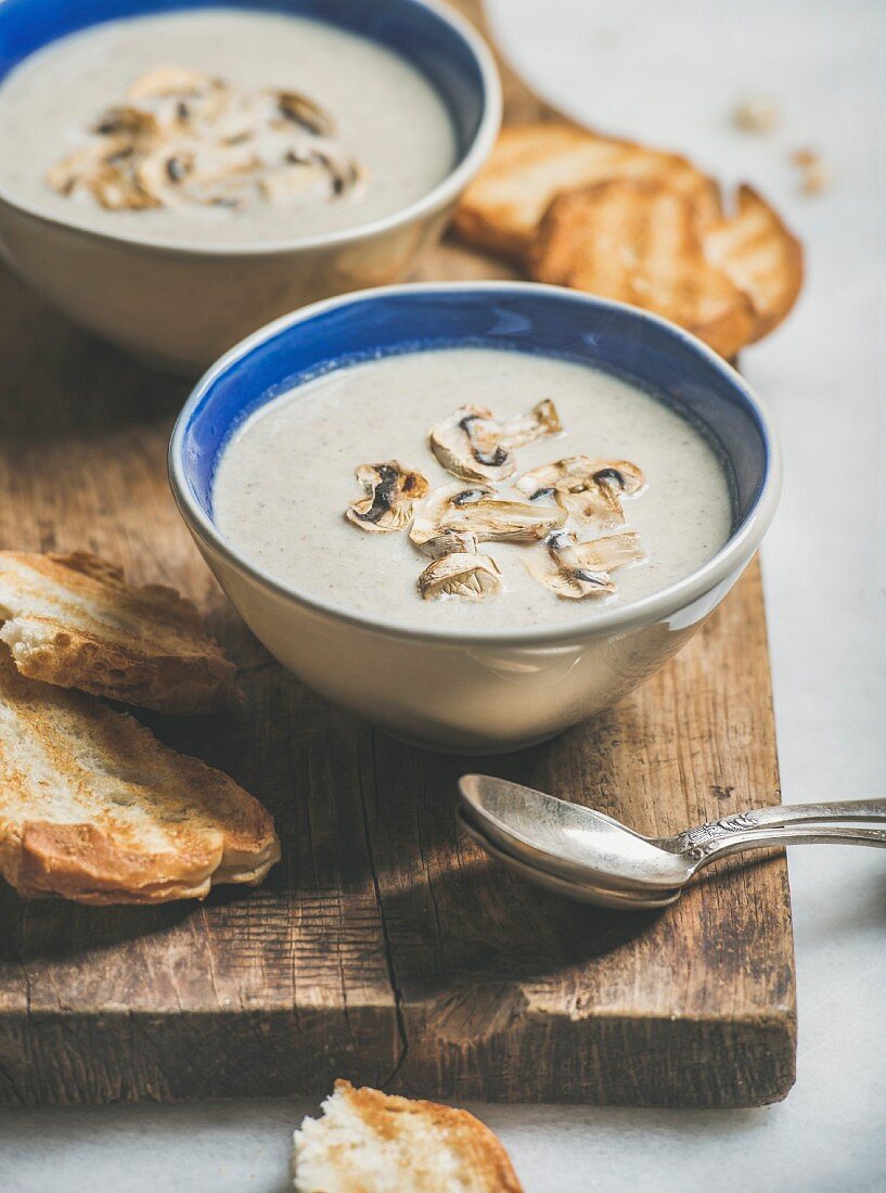 Pilzcremesuppe in Schälchen mit Röstbrot auf rustikalem Holzbrett