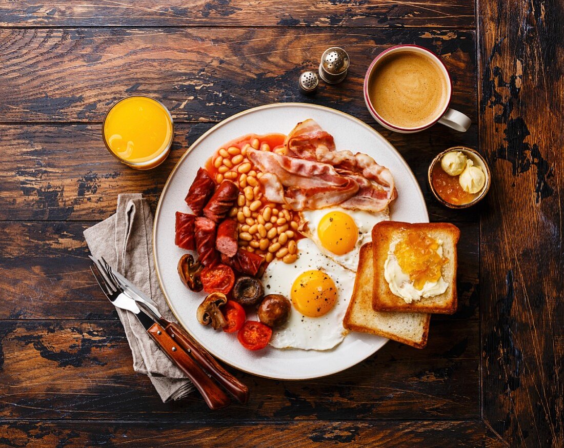 Full English breakfast with fried eggs, sausages, bacon, beans, toasts and coffee on wooden background